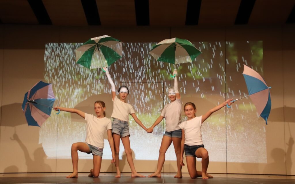Tanzaufführung mit vier Mädchen, die Regenschirme vor Regenprojektion halten.